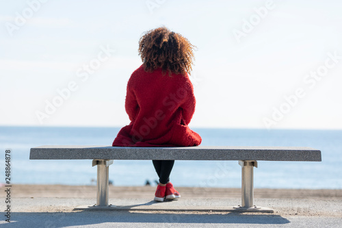Rear view of a young curly woman wearing red denim jacket sitting on a bench while looking away to horizon over sea