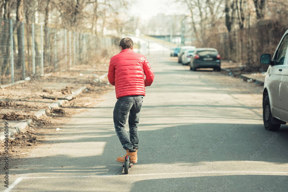 Fototapeta premium Young fashion guy riding electric scooter on the street, e-scooter kickscooter for personal transportation device in smart city wearing red jacket and working boots
