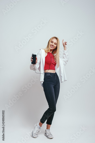 Favourite song. Full length portrait of beautiful young woman listening music with her smart phone, dancing and smiling while standing against grey background