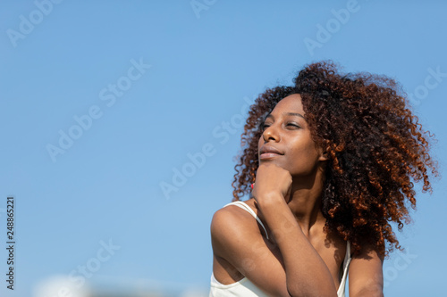 Front view of a young beautiful curly smiling woman wearing a white dress while sitting on ground and looking away in a sunny day