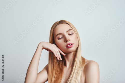 Perfect skin. Charming young woman with long blond hair touching her soft skin while standing against grey background. Skincare concept