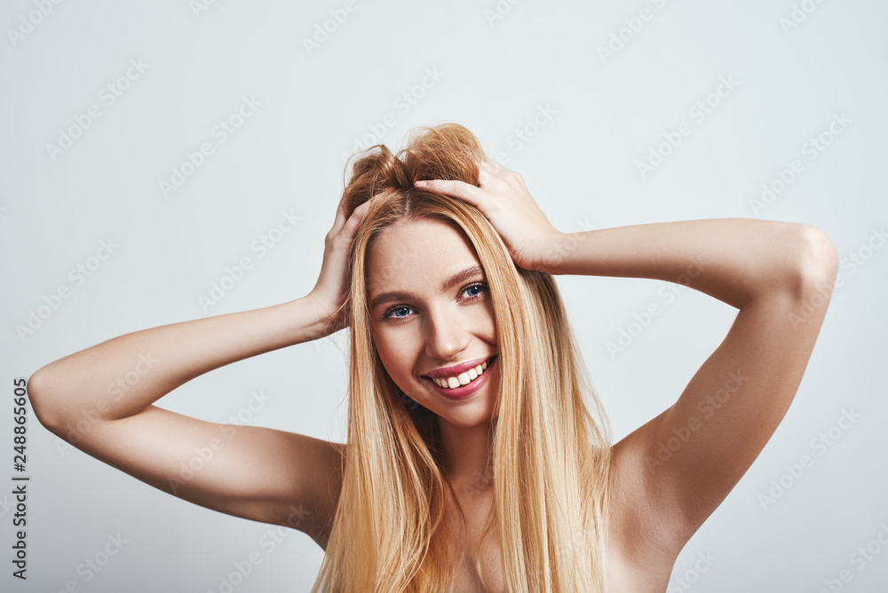 Studio shot of pretty smiling blonde woman playing with her hair and looking at camera standing on a grey background. Happiness concept