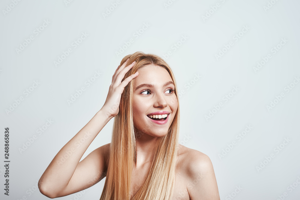Naklejka premium Feeling free and happy. Attractive young woman with naked shoulders touching her hair, looking away and smiling in studio on a grey background