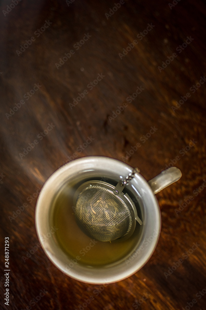 Cup of Tea with infuser in hot smoky water with green tea, mint herbs and honey on a dark wooden background Cup of Tea with infuser in hot smoky water with green tea, mint herbs and honey on a dark wooden background