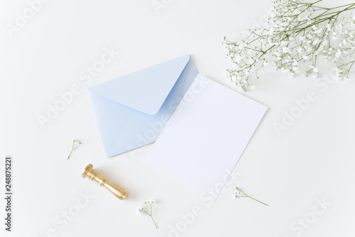 White and blue wedding stationery. Blank greeting card, craft envelope, washi tape and golden stamp, binder clips with olive branch.White table background. Flat lay, top view.