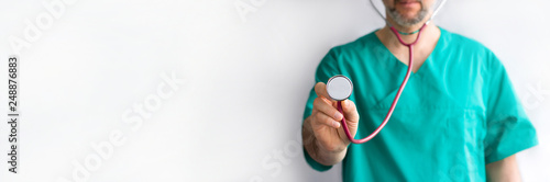 defocused doctor with beard in green uniform with stetoscope in hand in focus ready to examin on white background with copy space. Horizontal long banner, medicine, cardiology, heart checkup photo