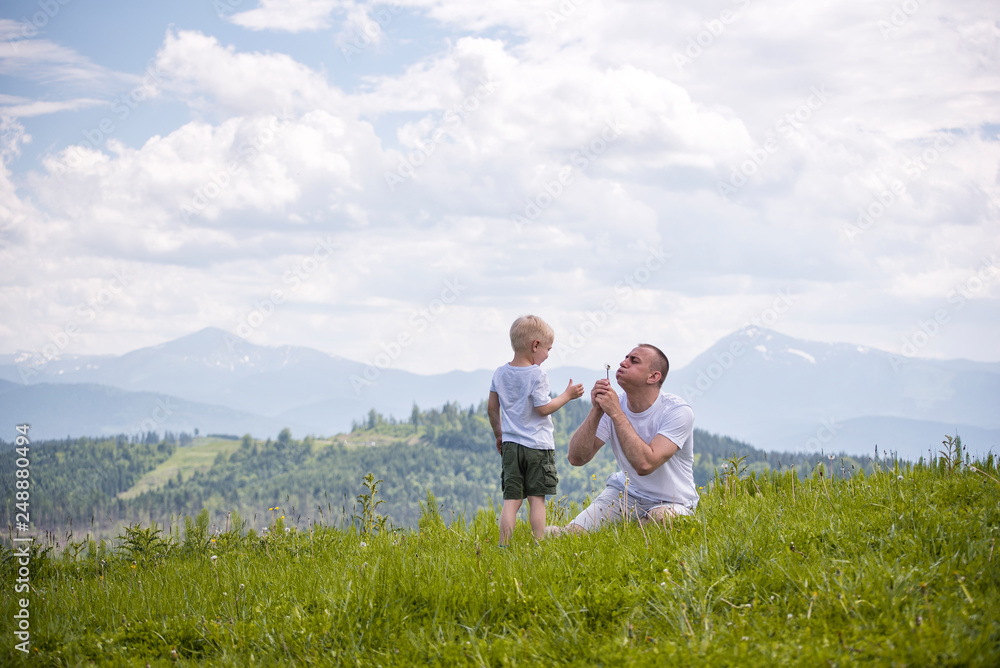 Father and young son are blowing dandelions sitting in the grass on a background of green forest, mountains and sky with clouds. Friendship concept Father and young son are blowing dandelions sitting in the grass on a background of green forest, mountains and sky with clouds. Friendship concept