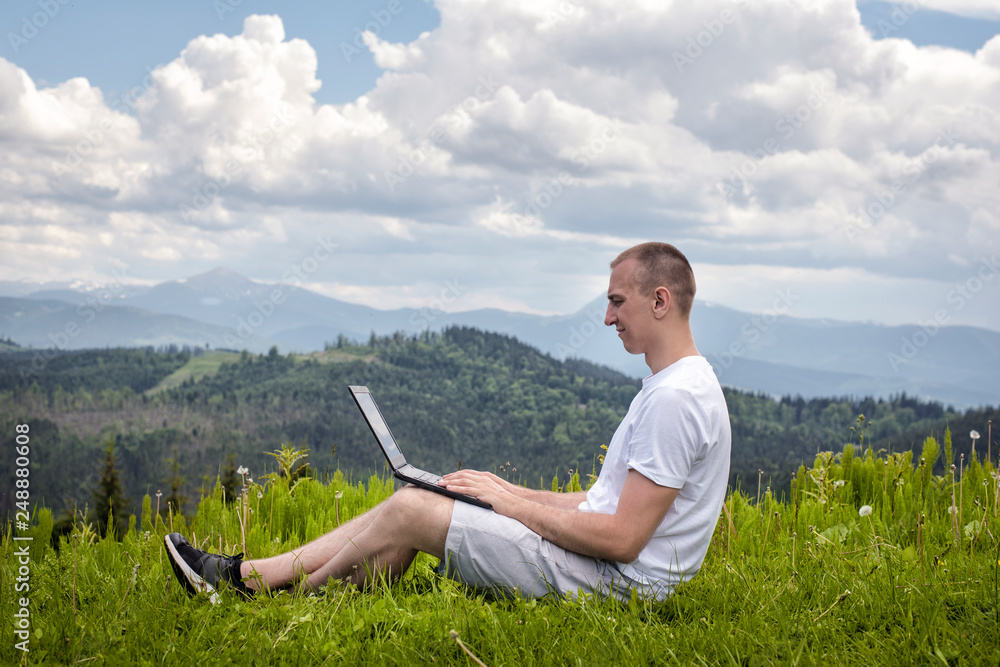 Man with laptop sitting on green grass on a background of mountains. Side view