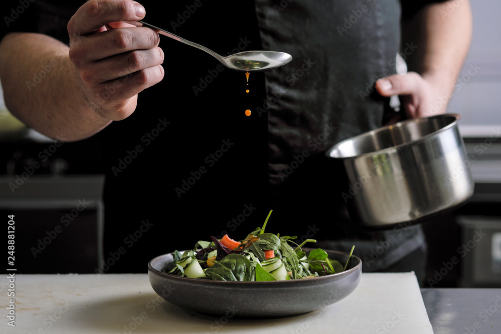 Close-up of the hands of a male chef on a black background. Pour sauce from the spoon on the salad dish. Close-up of the hands of a male chef on a black background. Pour sauce from the spoon on the salad dish.