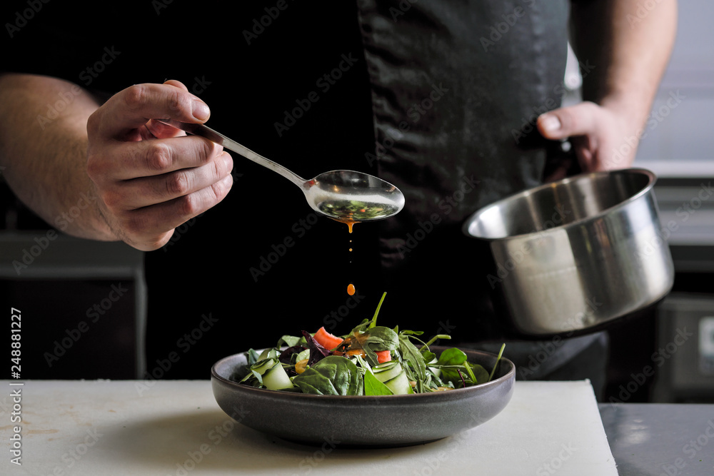 Close-up of the hands of a male chef on a black background. Pour sauce from the spoon on the salad dish. Close-up of the hands of a male chef on a black background. Pour sauce from the spoon on the salad dish.