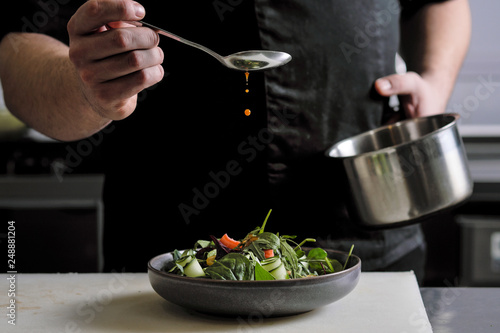 Close-up of the hands of a male chef on a black background. Pour sauce from the spoon on the salad dish.