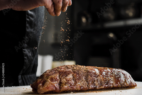 Raw piece of meat, beef ribs. The hand of a male chef puts salt and spices on a dark background.