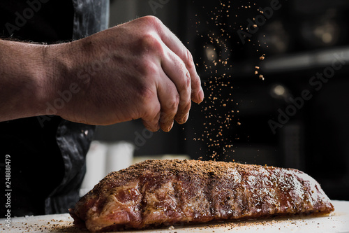 Raw piece of meat, beef ribs. The hand of a male chef puts salt and spices on a dark background.