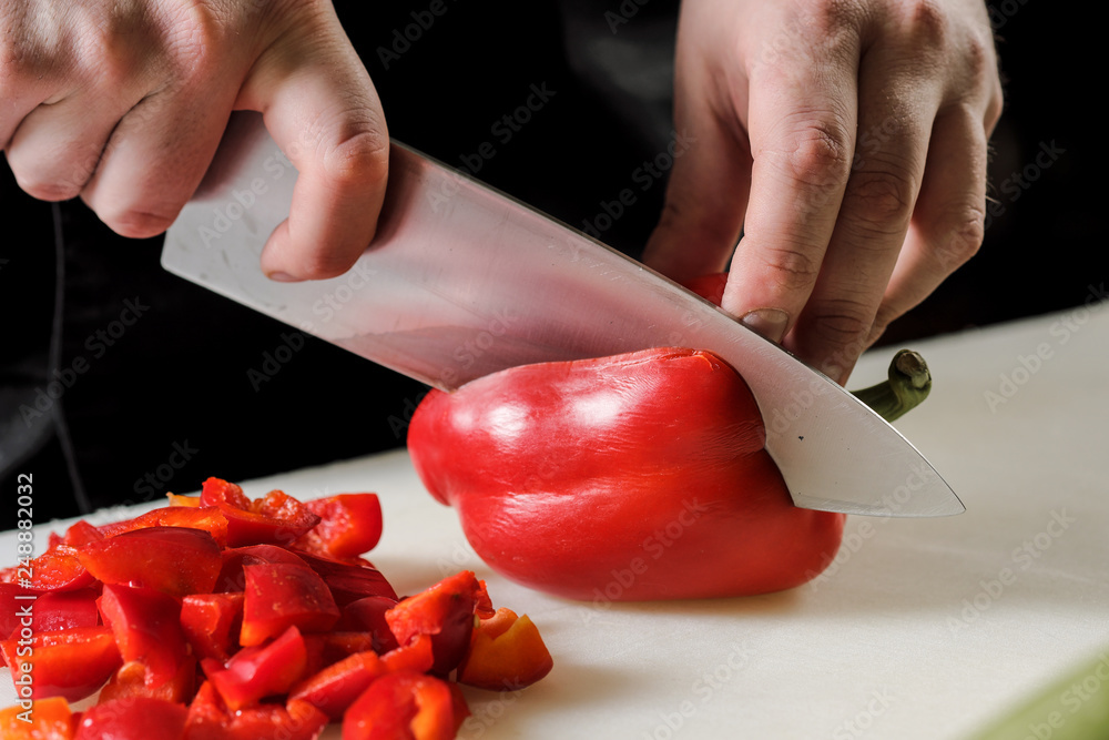 Male chef cuts vegetables for salad in a restaurant in a black apron. White cutting board, closeup of hands. Male chef cuts vegetables for salad in a restaurant in a black apron. White cutting board, closeup of hands.