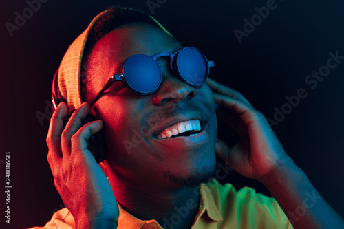 The young handsome happy hipster man listening music with headphones at black studio with neon lights. Disco, night club, hip hop style, positive emotions, face expression, dancing concept photo