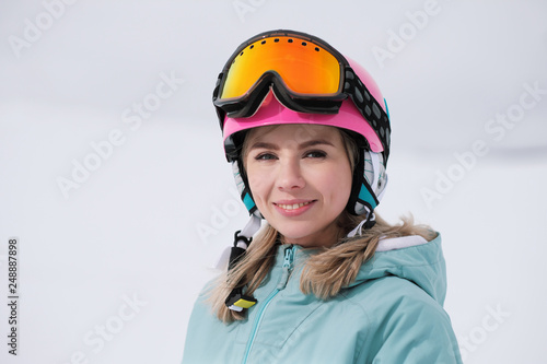 A beautiful young girl in an extreme sports suit, a hat and a helmet with a mask on the winter slope is holding alpine skis. © spaskov