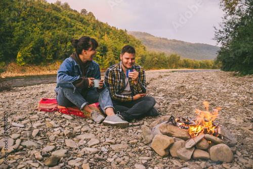 two young couple sitting near campfire drinking warm up tea and talking