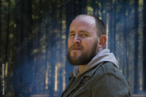Portrait of contented Adult young man in a tourist suit on nature in the spring forest on the background of tree trunks and blue smoke from a fire