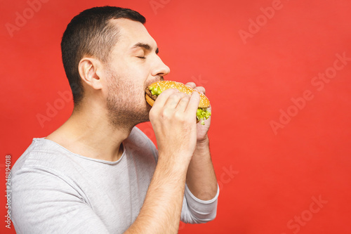 Young man holding a piece of hamburger. Student eats fast food. Burger is not helpful food. Very hungry guy. Diet concept. Isolated over red background.