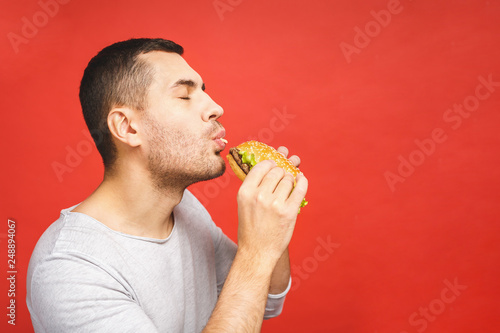 Young man holding a piece of hamburger. Student eats fast food. Burger is not helpful food. Very hungry guy. Diet concept. Isolated over red background.