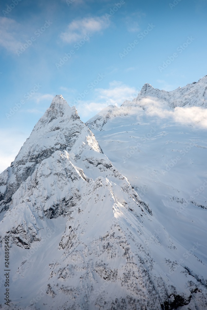 mountain peaks of Dombai mountains covered with snow surrounded by thick clouds against the blue sky. February 2019 mountain peaks of Dombai mountains covered with snow surrounded by thick clouds against the blue sky. February 2019