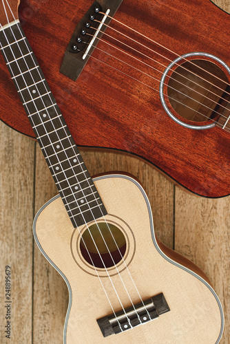 Guitar theme. Vertical top view of the acoustic and ukulele guitars lying on each other against the wooden floor.