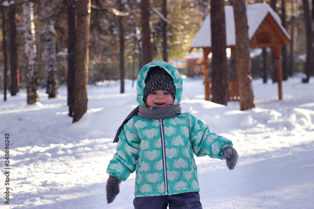 A blue-eyed little girl in a knitted hat, scarf and green winter jacket is laughing, rejoicing in the snow. Snow background. Close up. A blue-eyed little girl in a knitted hat, scarf and green winter jacket is laughing, rejoicing in the snow. Snow background. Close up.