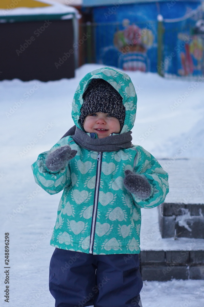 A blue-eyed little girl in a knitted hat, scarf and green winter jacket is laughing, rejoicing in the snow. Snow background. Close up.