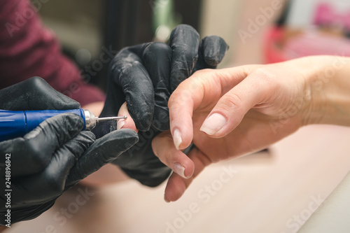 A manicurist in black latex gloves makes a hardware manicure to a client using a manual electric milling machine in a beauty salon  close up.