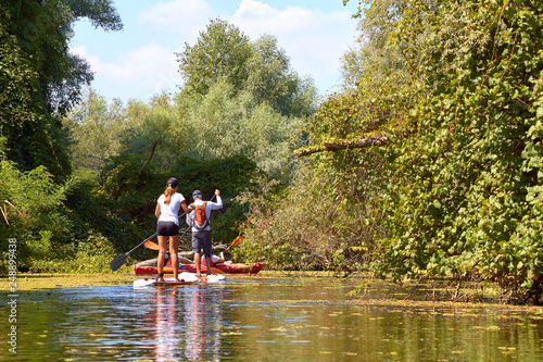 Athletic teen girl and man paddling on the SUP (stand up paddle board, paddleboard) in wilderness river overgrown duckweed near thickets of trees and wildgrapes at summer