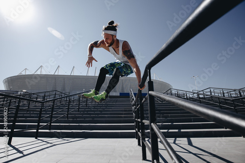 Young athletic man dressed in the white t-shirt, black leggings and blue shorts is jumping over the railing on the stairs in the street