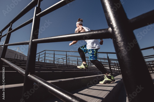Athletic guy with headband on his head dressed in the white t-shirt, black leggings and blue shorts is running up the stairs with black railings outside on a sunny day