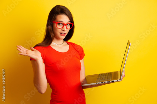 Beautiful student in red tshirt and red glasses holds a laptop over yellow background