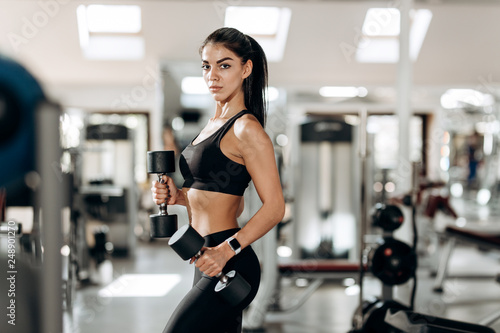 Athletic girl dressed in black sports top and tights builds up muscles with dumbbells in the gym
