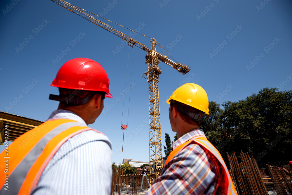 Two men dressed in orange work vests and helmets look at th crane on the building site Two men dressed in orange work vests and helmets look at th crane on the building site