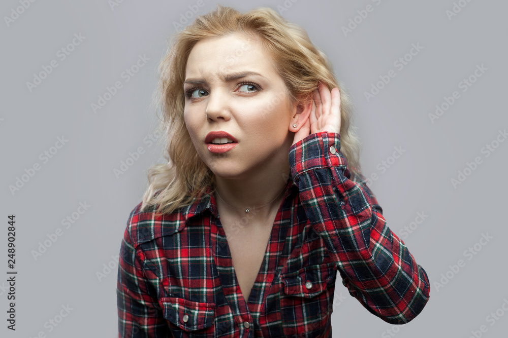 Portrait of nosy beautiful blonde young woman in casual red checkered shirt standing with hand near ear and trying to hear and spying. indoor studio shot, isolated on grey background. Portrait of nosy beautiful blonde young woman in casual red checkered shirt standing with hand near ear and trying to hear and spying. indoor studio shot, isolated on grey background.
