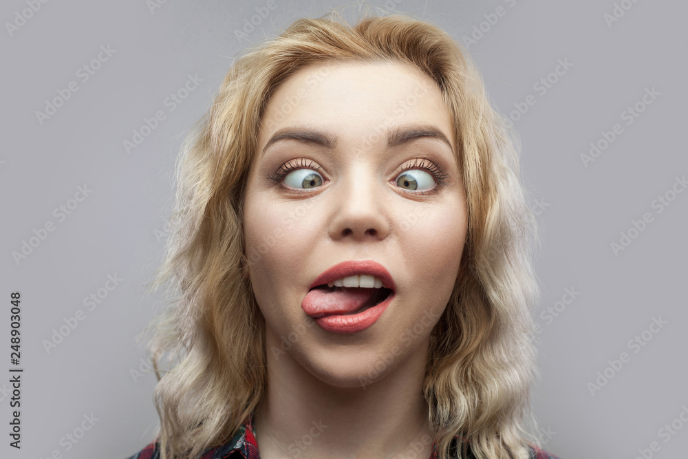 Portrait of funny crazy young beautiful blonde woman in casual red  checkered shirt standing and looking with crossed eyes and tongue out.  indoor studio shot, isolated on grey background. Stock Photo |