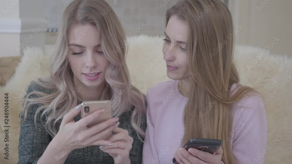 Two beautiful young girlfriends sitting on the couch in the living room looking for good discounts for shopping using internet in their gadgets. Shopping concept.