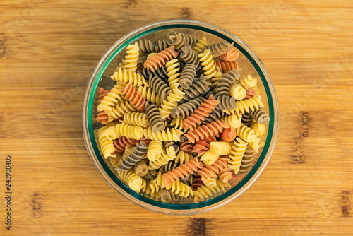 Raw uncooked three-colored Fusilli in a glass bowl on wood table.