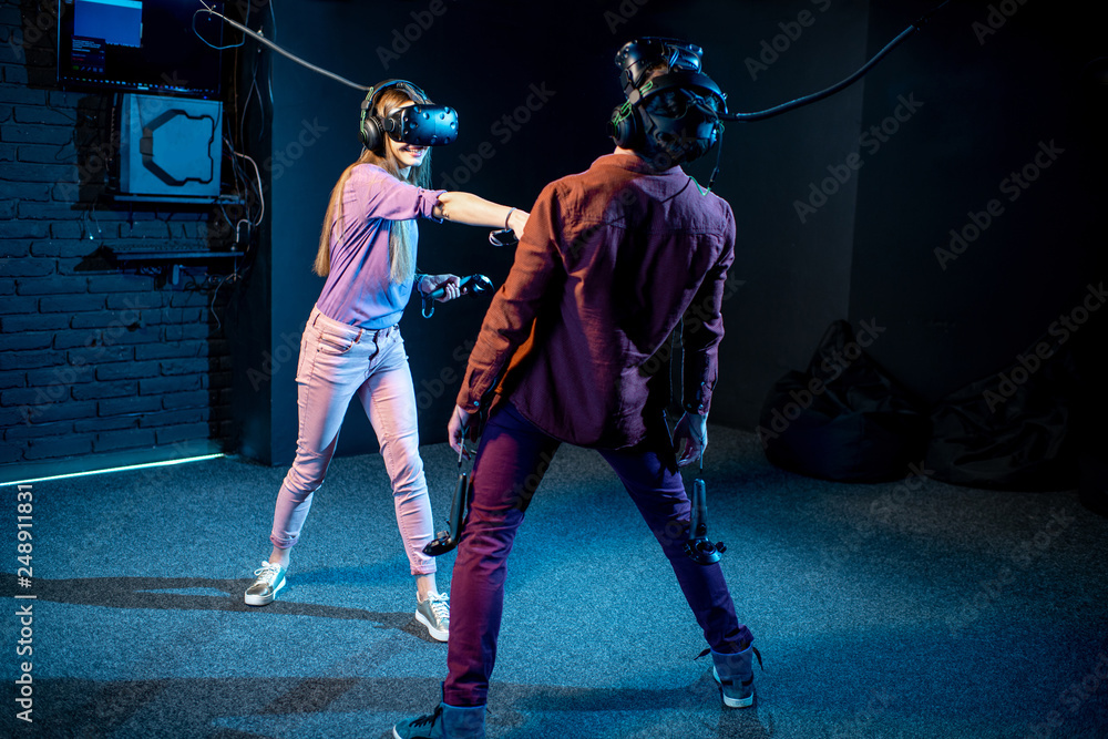 Fototapeta premium Man and woman playing game using virtual reality headset and gamepads in the dark room of the playing club