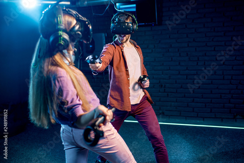 Man and woman playing game using virtual reality headset and gamepads in the dark room of the playing club