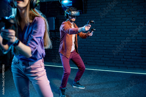 Man and woman playing game using virtual reality headset and gamepads in the dark room of the playing club