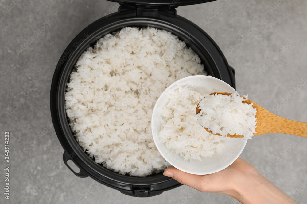 Woman putting boiled rice into bowl from cooker on grey background, top view Woman putting boiled rice into bowl from cooker on grey background, top view