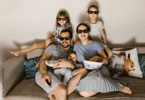 Happy family father with baby on his arms, mother and two daughters in the special glasses watching tv and eating popcorn sitting on the sofa