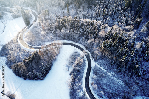 Aerial drone view of a curved winding road through the forest up in the mountains in the winter with snow covered trees and curved streets in winter while sunset and sun rays casting trough © visualitte