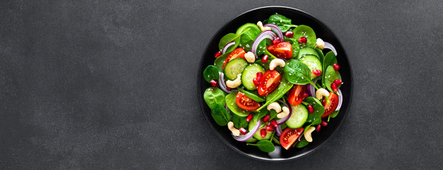 Spinach salad with fresh cucumbers, tomato, onion, pomegranate, sesame seeds and cashew nuts on black background. Healthy vegan food. Top view. Banner