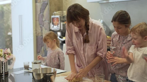 Mother takes huge piece of dough and tries to show her kids how to knead a dough. They are excited to learn and help. Eldest daughter brings a glass for making circles and baking them into cookies. photo