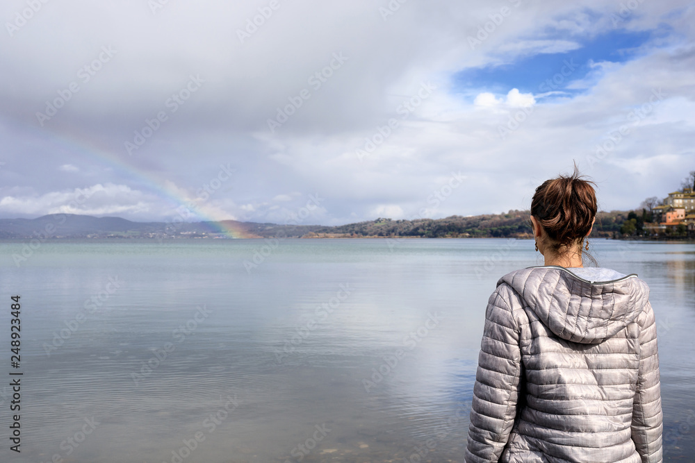 Girl at the lake and rainbow. Girl on the shore of the lake looks at the rainbow on the horizon. On a winter morning at Lake Bracciano in Italy.