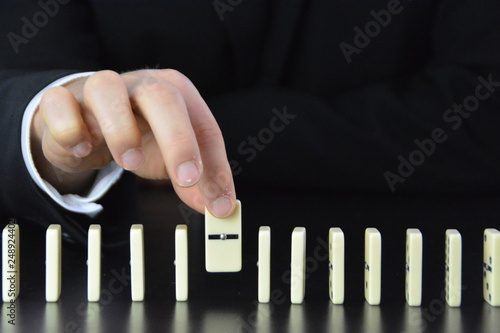 A young man in a suit and jacket removes a domino from a series of dominoes and prevents such a chain reaction when falling over - concept with a business man who intervenes before a catastrophe