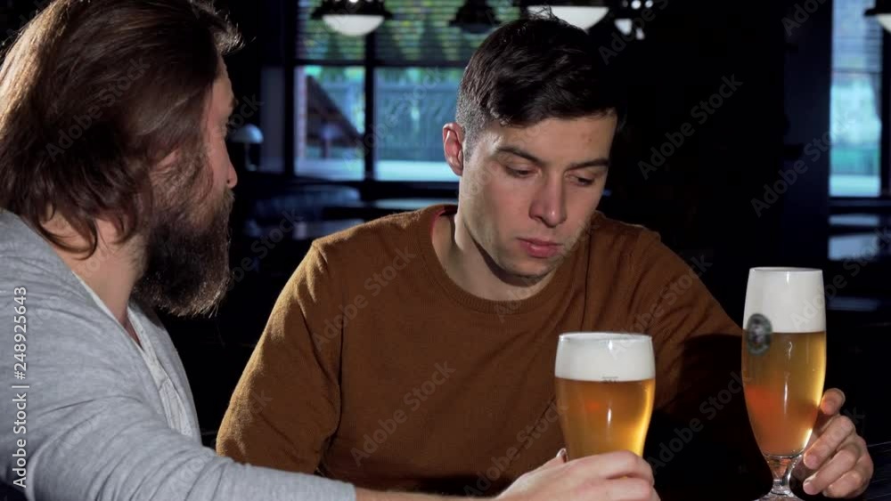 Depressed young man drinking at the bar with his friend. Handsome man looking upset, clinking glasses with a friend, drinking beer at the pub. Bearded man supporting his sad friend Depressed young man drinking at the bar with his friend. Handsome man looking upset, clinking glasses with a friend, drinking beer at the pub. Bearded man supporting his sad friend